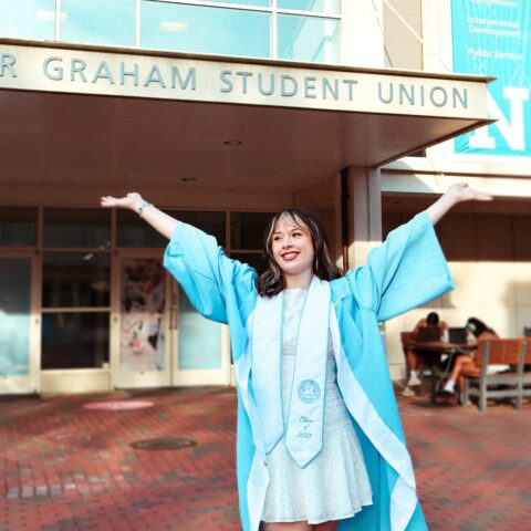 Student staff member Savannah Nelson poses in from of the Union in graduation attire for grad photos.