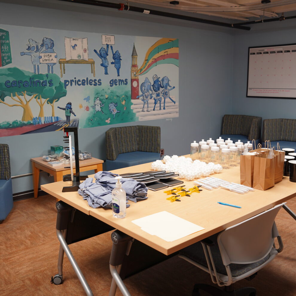 A room with a desk, chairs, and a mural on the wall. On the desk there are bottles, papers, pens, and other objects. There are also sofas and tables in the room.