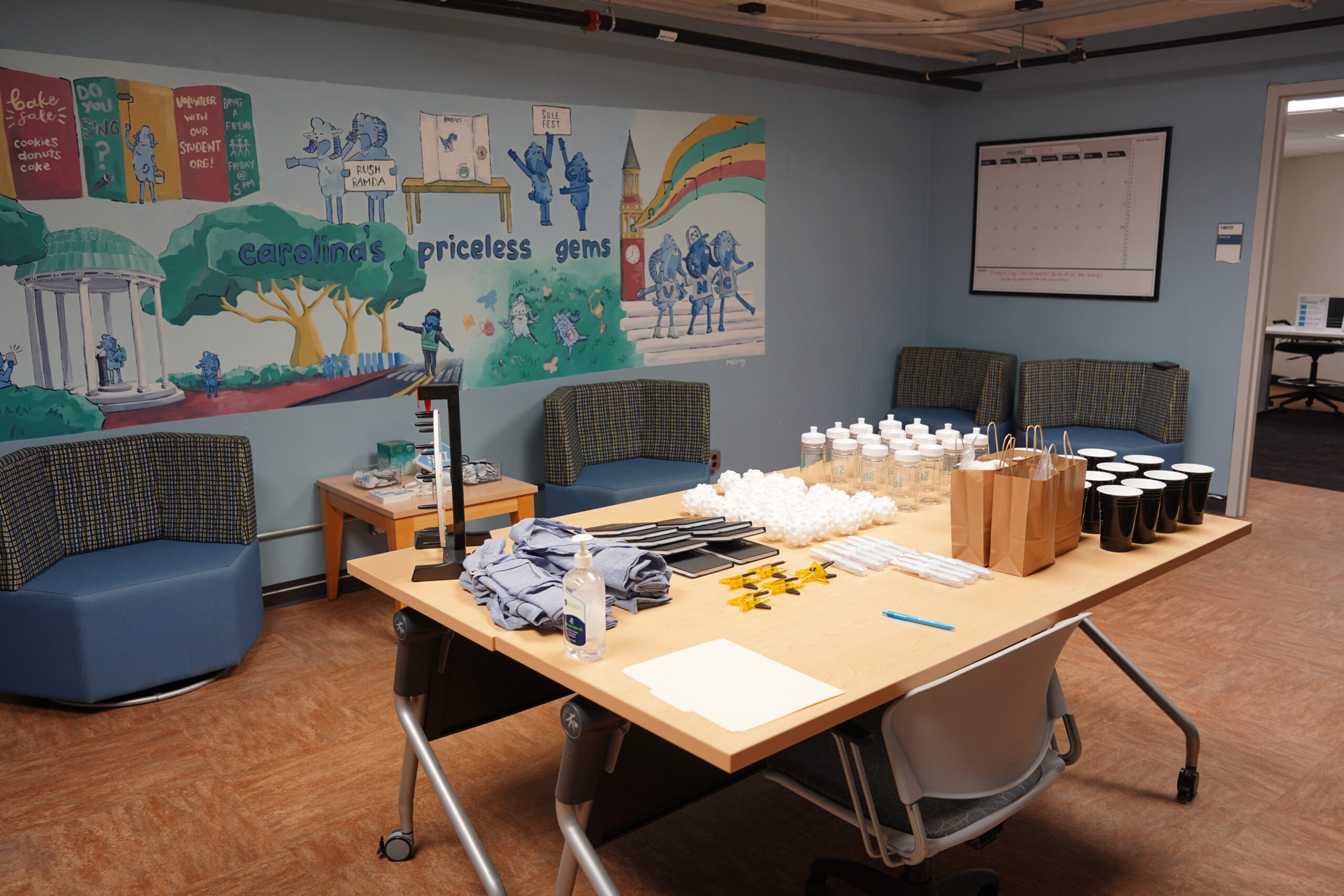 A room with a desk, chairs, and a mural on the wall. On the desk there are bottles, papers, pens, and other objects. There are also sofas and tables in the room.