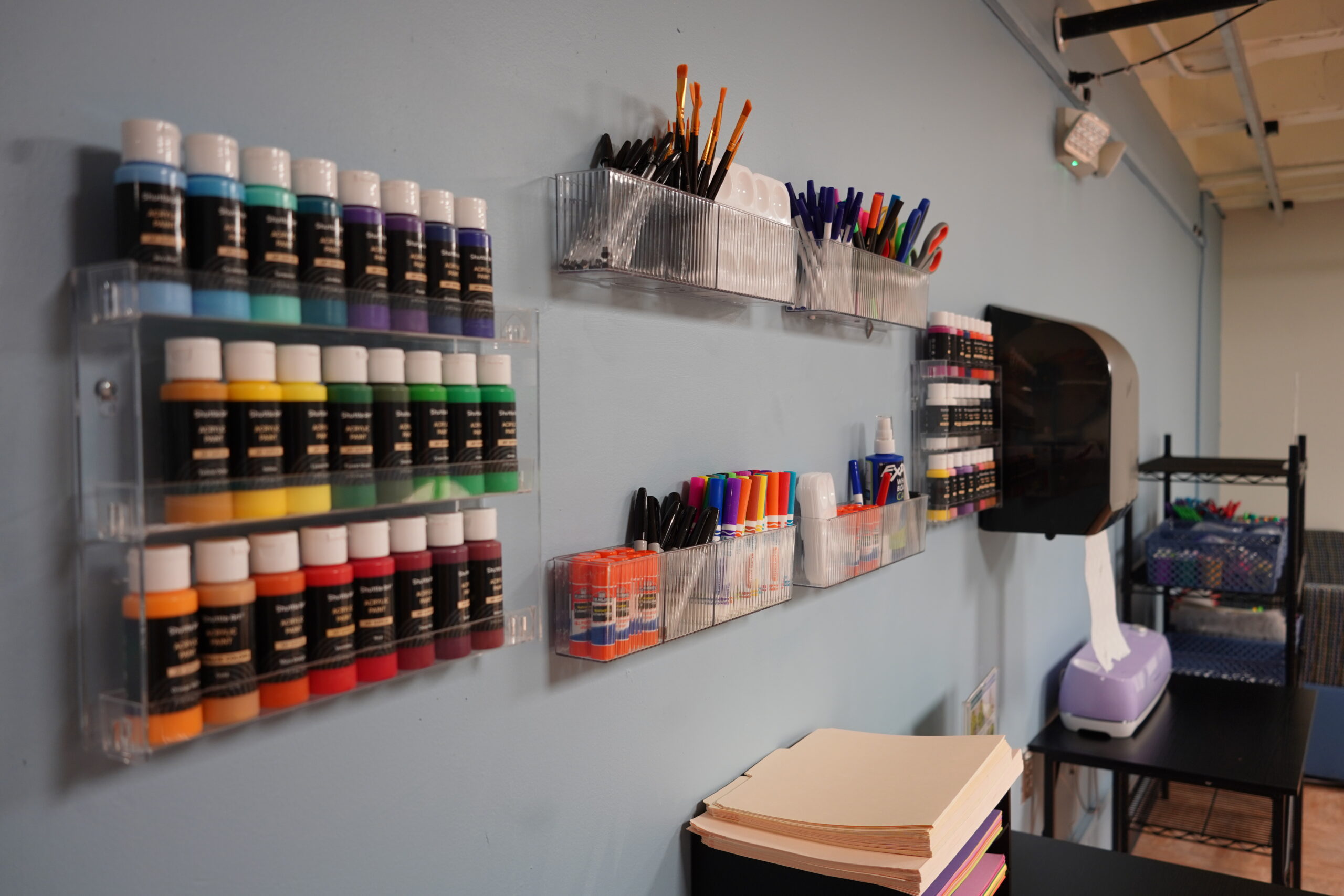 A wall with organized craft supplies, including paint bottles in various colors, pens, and markers. Nearby are stacks of paper and a shelving unit with additional office items.