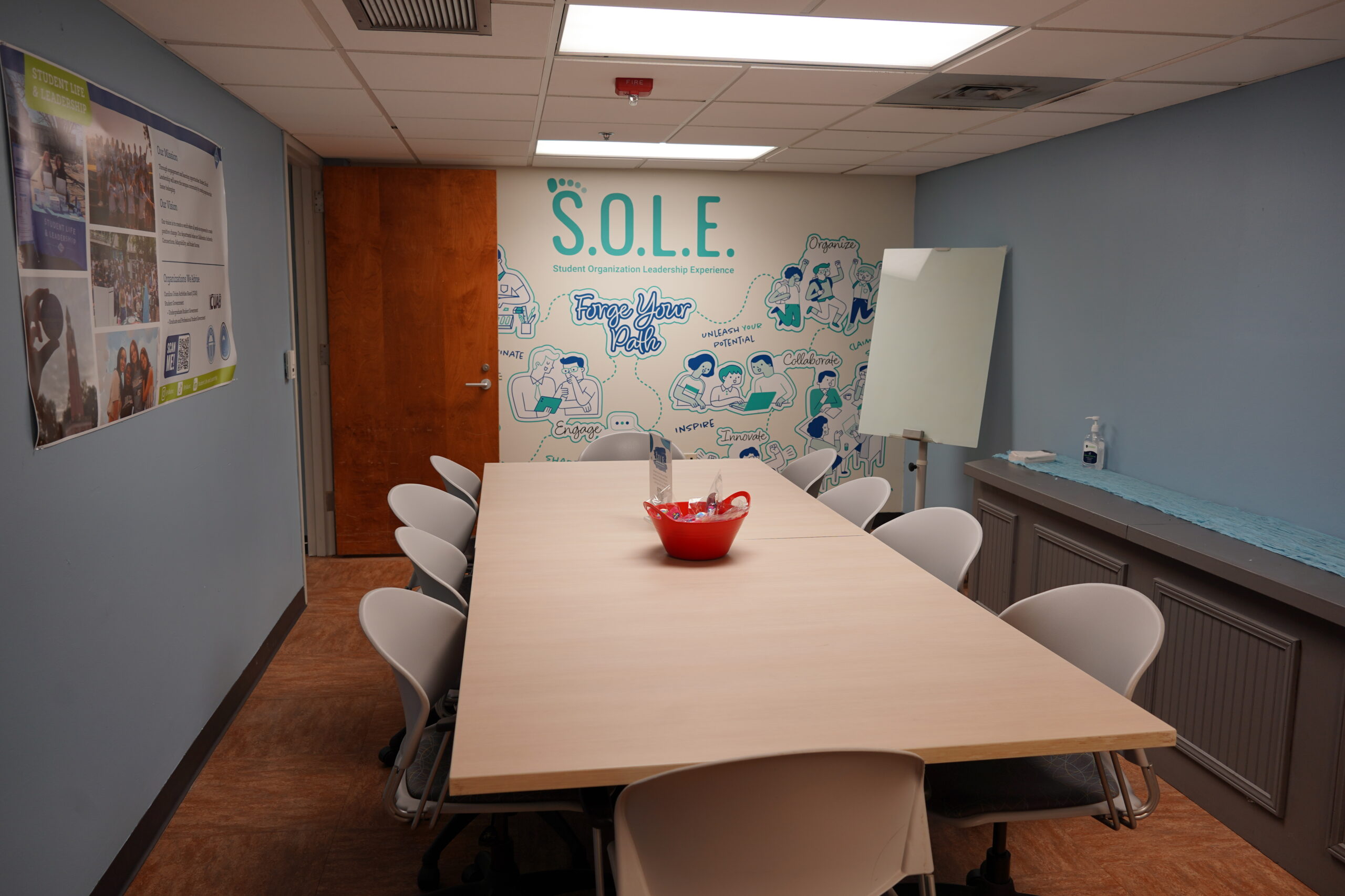 A conference room with a rectangular table surrounded by nine white chairs. The room has a light blue wall with the "S.O.L.E." logo and illustrations.
