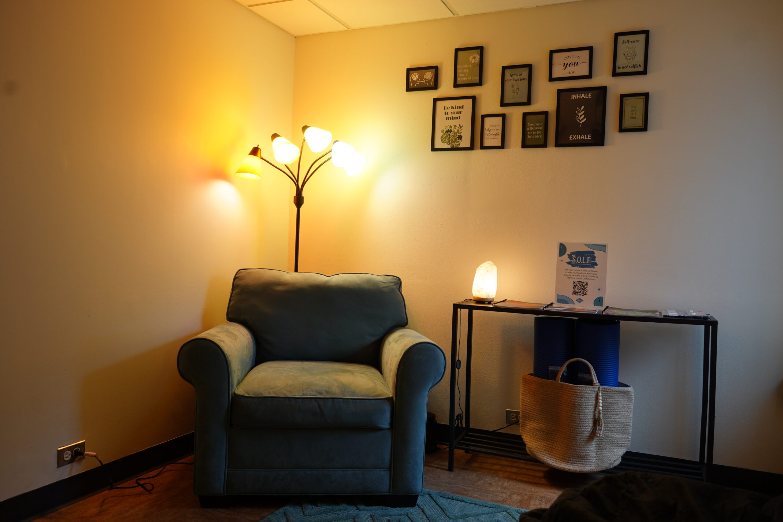 A cozy room featuring a plush armchair under a floor lamp with multiple lights. The wall is adorned with framed inspirational quotes. A small table holds a salt lamp. A woven basket sits below the table.